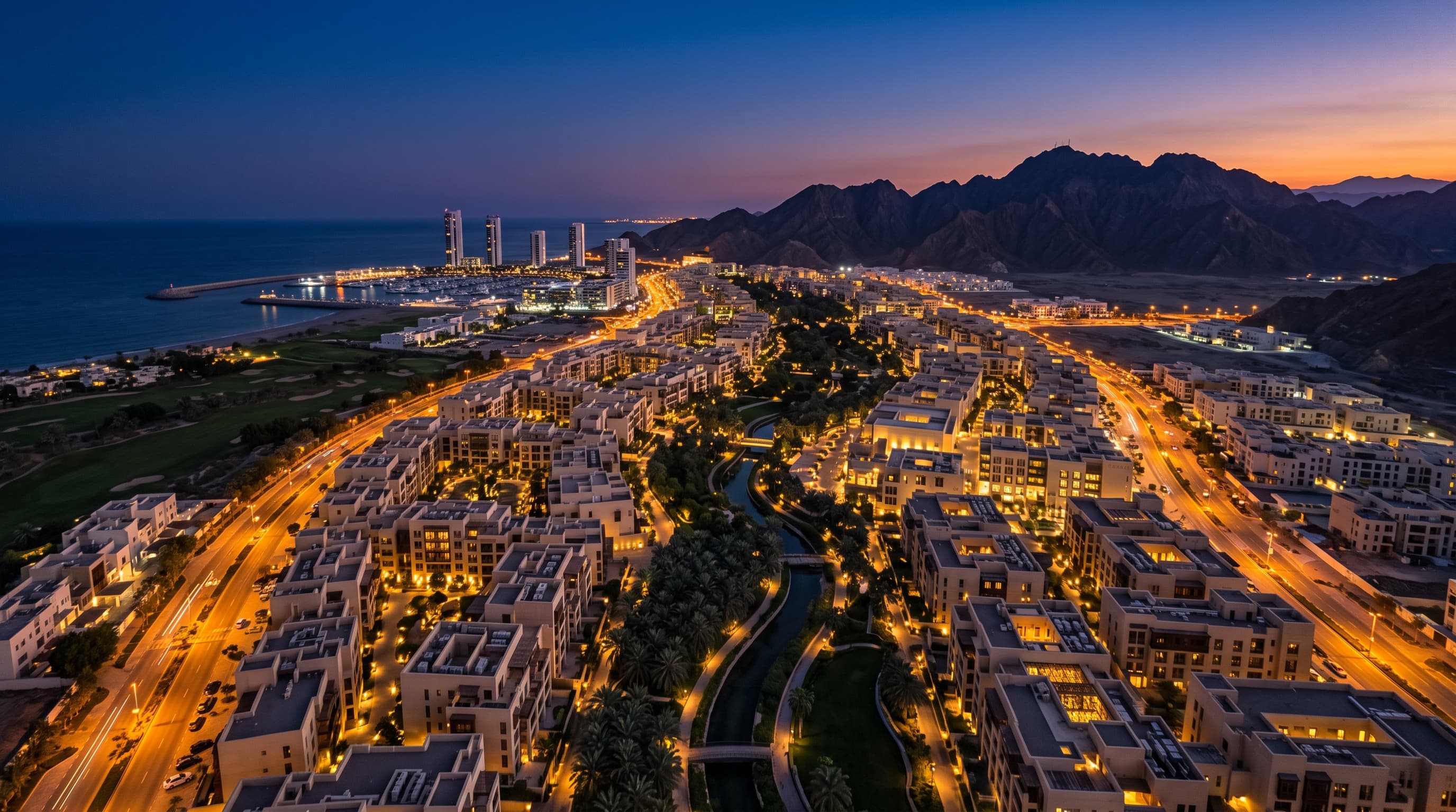 Aerial view of Madinat Al Irfan at blue hour, with the wadi park winding through the city and mountains beyond